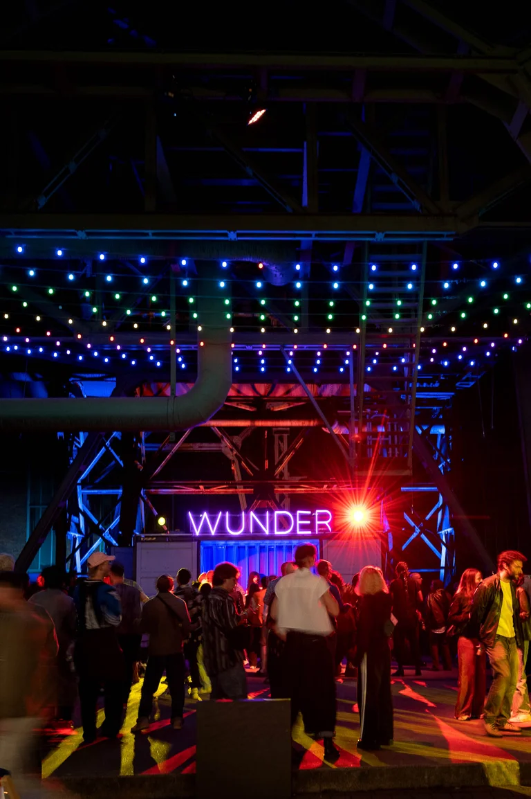Eine Menschenmenge tanzt vor einer dunklen Industriehalle unter Reihen bunter Lichterketten. Im Hintergrund leuchtet der Schriftzug „WUNDER“ in violettem Neonlicht über einem blau beleuchteten Container, daneben strahlt ein roter Scheinwerfer.