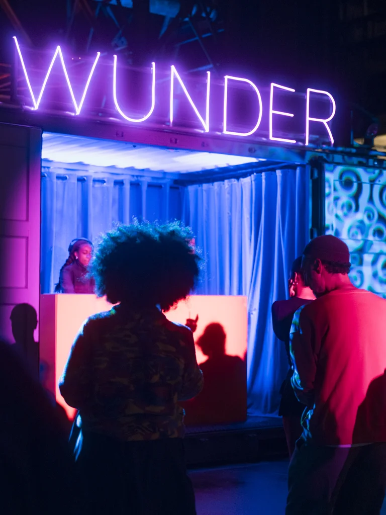 Eine DJ legt in einem blau und pink beleuchteten Container auf, über dem der Schriftzug „WUNDER“ in violettem Neonlicht leuchtet. Im Vordergrund tanzen mehrere Personen im farbigen Licht.