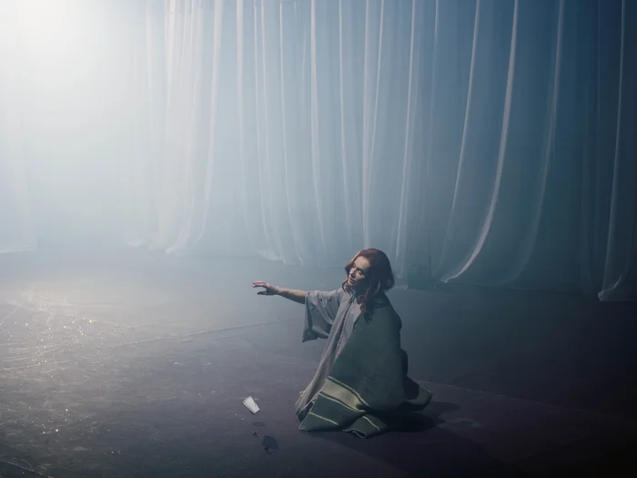 Eine Frau in einem Kleid, die Schauspielerin Isabelle Huppert, sitzt vor einem Vorhang auf dem Boden