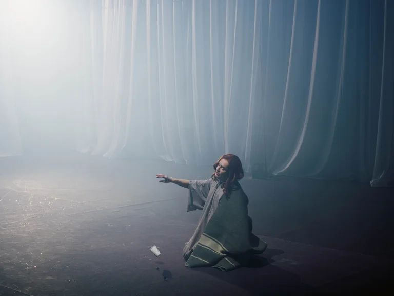 A woman in a dress, the actress Isabelle Huppert, is sitting on the floor in front of a curtain