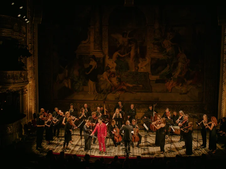Ein klassisches Orchester spielt auf einer prunkvollen Theaterbühne. Die Musikerinnen und Musiker sind in schwarzer Konzertkleidung gekleidet und spielen Streichinstrumente. In der Mitte steht der Sänger Samuel Mariño in einem auffälligen roten Anzug, während der Dirigent das Ensemble leitet.