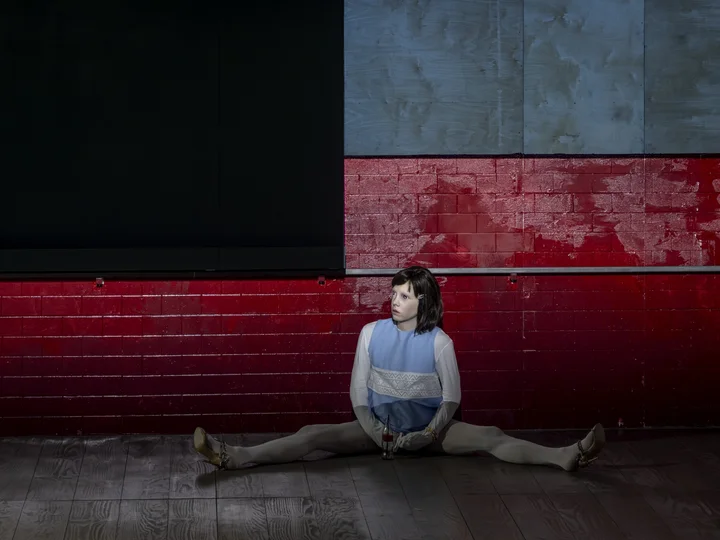 A person is sitting on the floor in front of a red wall with their legs stretched out. They are wearing a childlike mask.