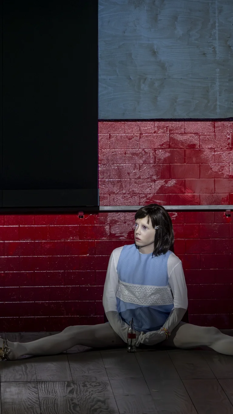 Eine Person sitzt mit weit ausgestreckten Beinen auf dem Boden vor einer roten Wand. Sie trägt eine kindlich anmutende Maske.