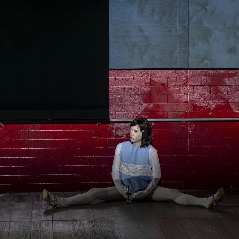 Eine Person sitzt mit weit ausgestreckten Beinen auf dem Boden vor einer roten Wand. Sie trägt eine kindlich anmutende Maske.