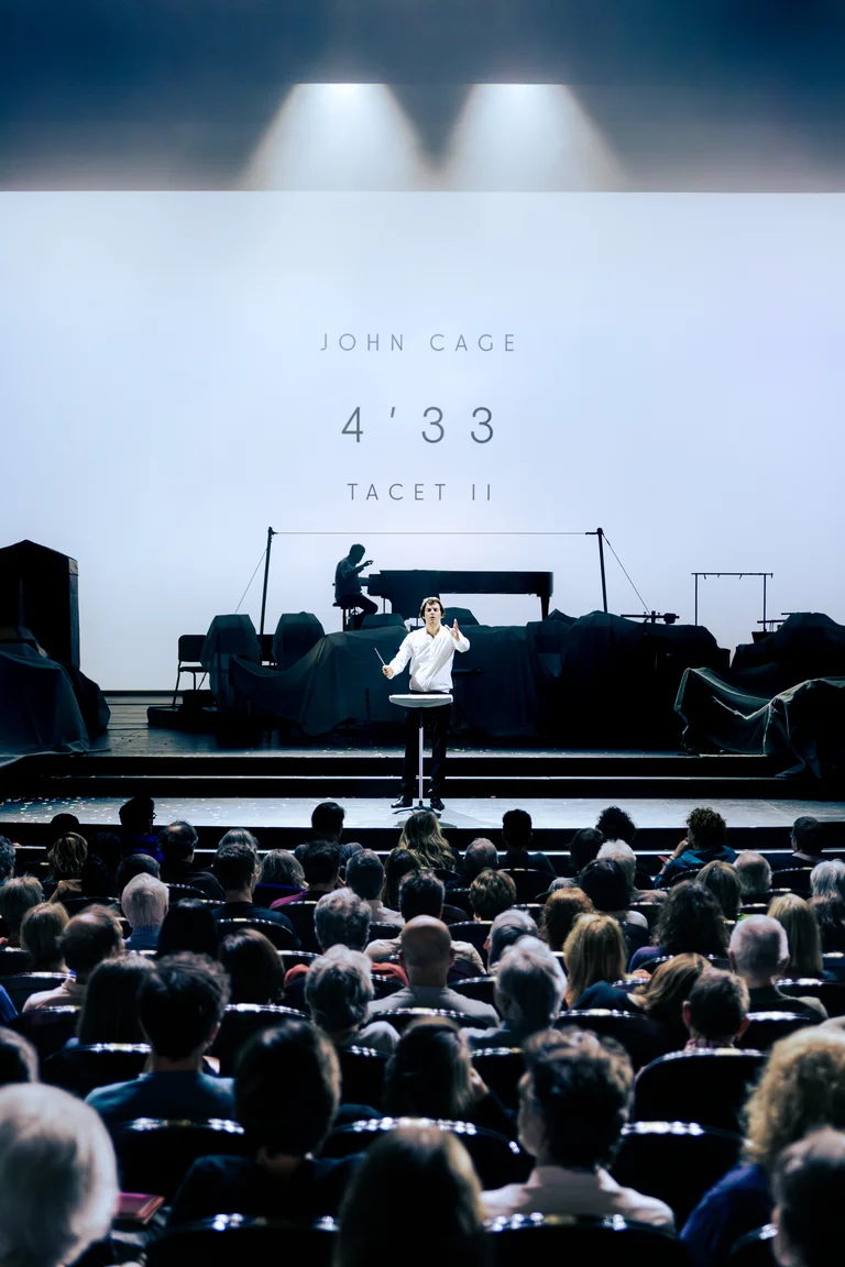 The ensemble Les Apaches ! performs on a stage in front of an audience. In the foreground, a conductor stands in the spotlight, while a pianist sits at a grand piano in the background. The words “John Cage 4’33 Tacet II” are displayed on a screen.