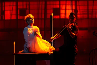 Joyce DiDonato sits on a wooden table in a white dress, holding one hand to her chest. Beside her, Nick Kendall plays the violin. The stage is bathed in intense red and orange light.