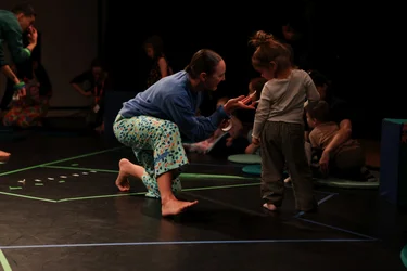 Eine Performerin interagiert mit einem kleinen Kind, während andere Kinder im Hintergrund spielen. Der Boden ist mit bunten Linien markiert.