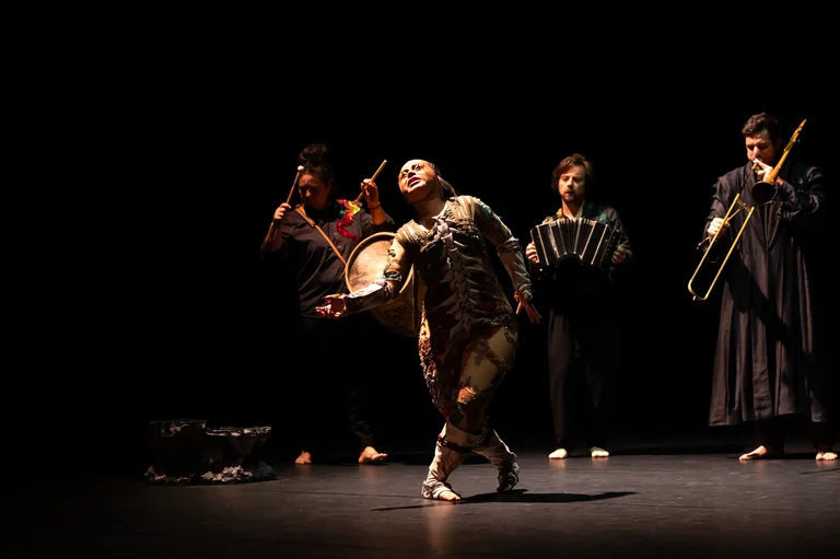In the foreground, a person dances in an elaborate costume with an expressive gesture; in the background, three musicians play drum, bandoneon, and trombone.