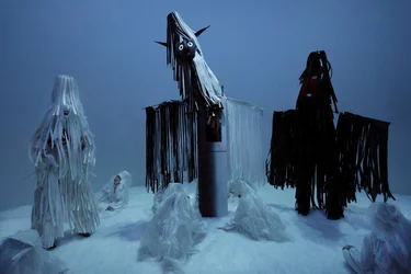 Scene from Balkan Erotic Epic: Several white-clad figures sit and stand in a snow-covered setting around a tall black figure with long fringes. The atmosphere feels ritualistic and mysterious.