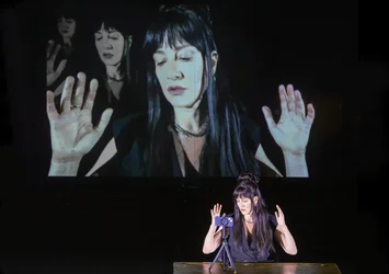 Eine Person sitzt an einem Tisch und spricht mit erhobenen Händen in ein Smartphone auf einem Stativ. Hinter ihr ist eine großformatige Video-Projektion von ihr selbst zu sehen.