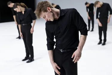 A performer in black clothing tilts their head downward while others in the background hold similar poses.