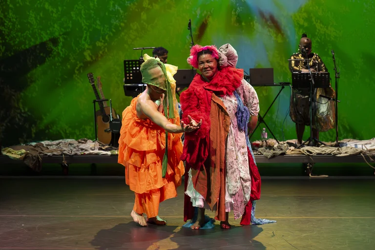 Two performers in colorful costumes, one in orange and one in red, stand on stage with musicians and green projections in the background.