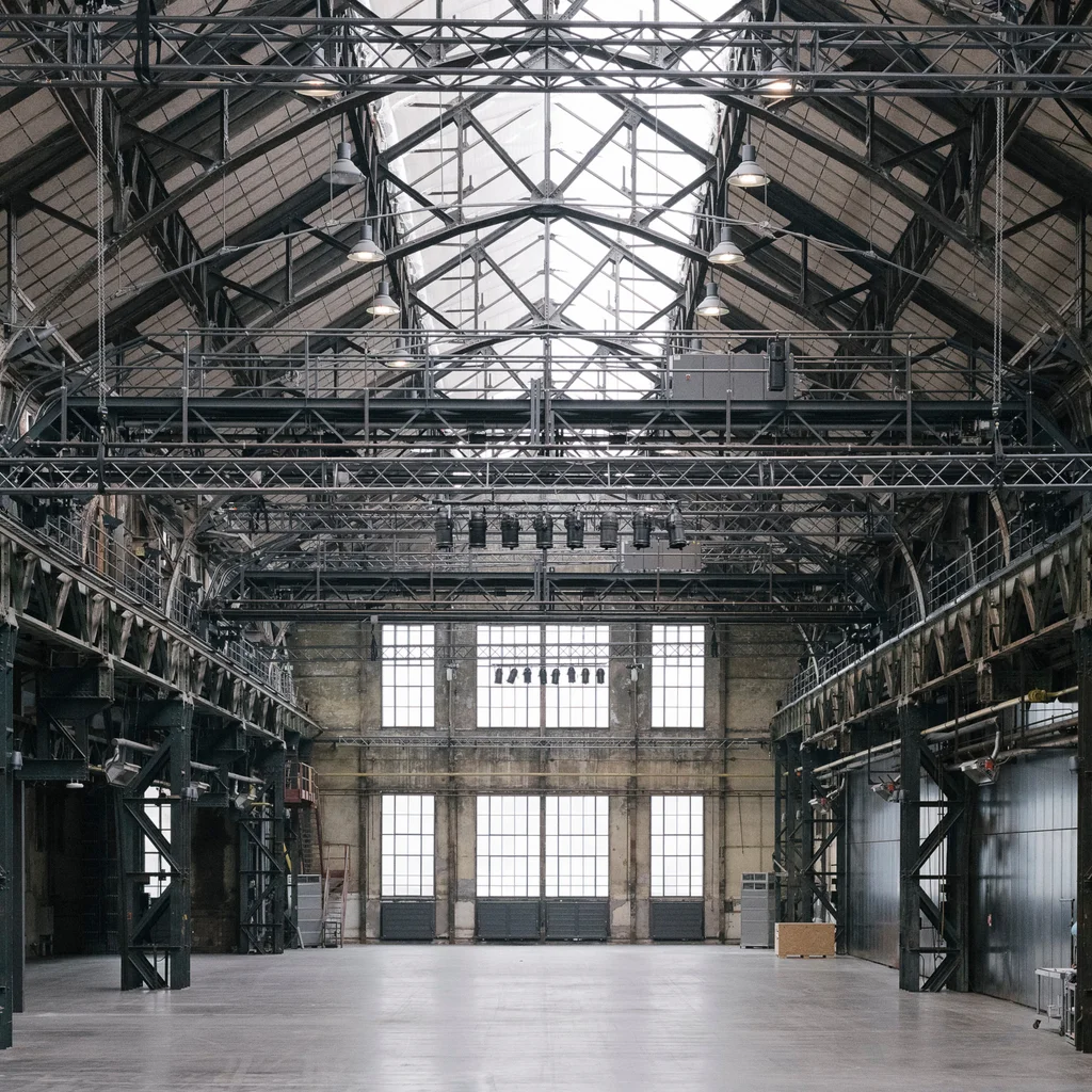 View of the interior of the Jahrhunderthalle Bochum, featuring its expansive space, high steel beams, and large windows letting in natural light. The hall appears both open and industrial.
