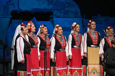 Eine Gruppe von Frauen singt auf einer Bühne vor Mikrofonen. Sie tragen traditionelle, reich verzierte Trachten mit floralen Kopfbedeckungen, bestickten Gewändern und auffälligem Silberschmuck.