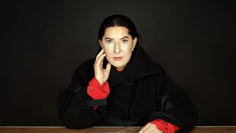 Portrait von Marina Abramović, die an einem Tisch sitzt und mit eine Hand ihr Gesicht berührt, die andere ruht auf dem Tisch neben einem weißen Blatt Papier.