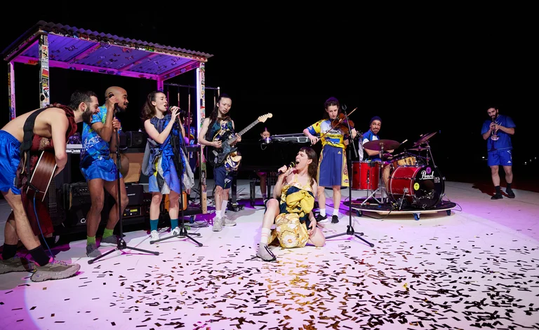 Tänzerinnen der Dresden Frankfurt Dance Company stehen im Kreis und formieren eine Band,  eine Sängerin kniet neben einem Schlagzeug. Auf dem Boden liegt goldenes Konfetti.