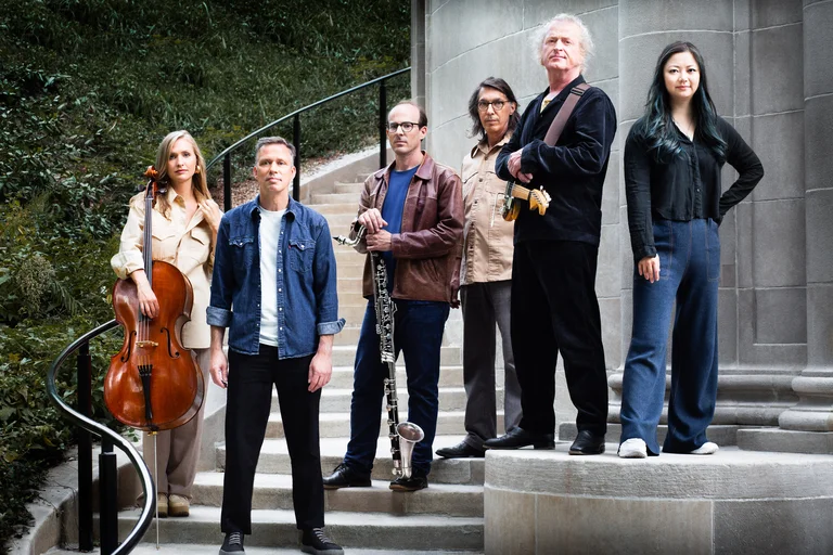 Die sechs Orchester-Mitglieder von Bang on a Can stehen mit ihren Instrumenten nebeneinander auf einer Steintreppe draußen vor einem Gebäude aufgereiht. Im Hintergrund sind grüne Sträucher zu sehen.