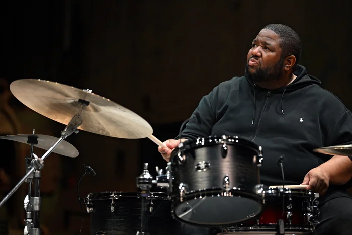 Drummer Tyshawn Sorey plays intently at the drum set. He is wearing a black hoodie and looks upward to the right. Cymbals and drums are visible in the foreground.
