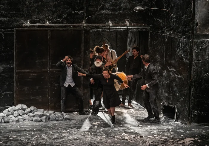 A theatre ensemble performs on a dark stage, with musicians, actors, and a woman in the foreground rushing forward with energy. Surrounded by stones and a broken stage setting, the scene conveys chaos and intensity.