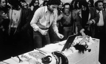 People are gathering in front of a table with various objects on it. The photograph was taken during Marina Abramović’s performance “Rhythm 0”.