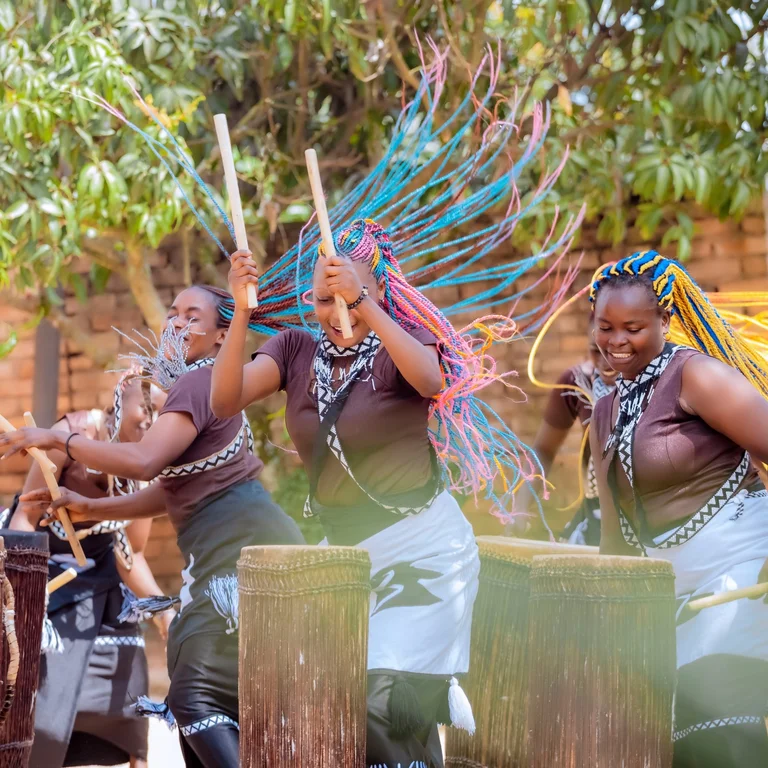 Drei Trommlerinnen schlagen auf ihre Instrumente, während Bewegung, Rhythmus und Gruppendynamik die Szene prägen.