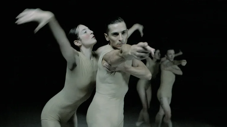 Two dancers in skin-toned costumes stand in the foreground, moving powerfully forward. In the background, additional dancers perform, enhancing the dynamism of the scene.