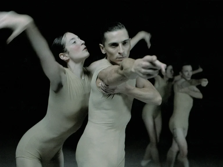 Zwei Tänzer*innen in hautfarbenen Kostümen stehen im Vordergrund und bewegen sich kraftvoll nach vorn. Im Hintergrund tanzen weitere Tänzer*innen und verstärken die Dynamik der Szene.