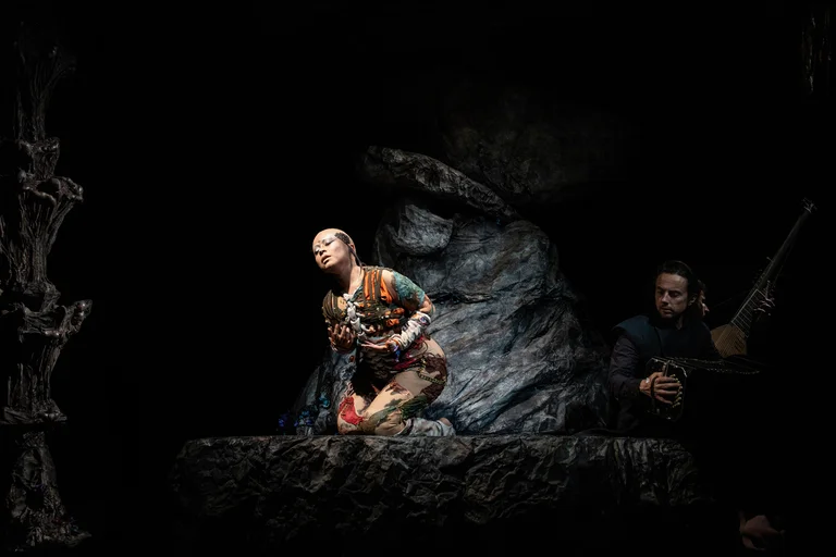 A woman kneels in a colorful costume on a rocky stage in front of an illuminated rock backdrop. On the right side of the image, a musician is seated, playing both a bandoneon and a theorbo. The background is dark.