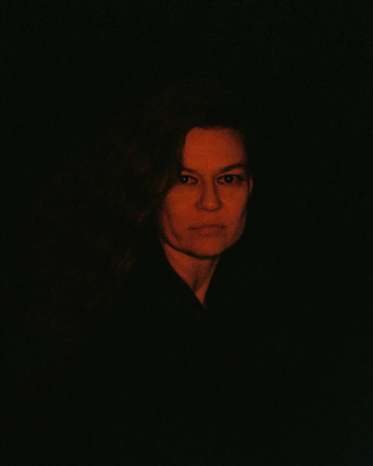 Sharon Eyal looks directly into the camera. Her face is illuminated by a warm, reddish light, while the background and her clothing are almost entirely shrouded in darkness.