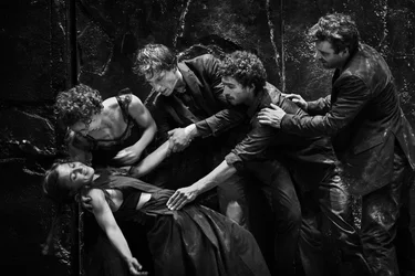Five performers in dark clothing enact a dramatic scene on stage. The photo is in black and white.