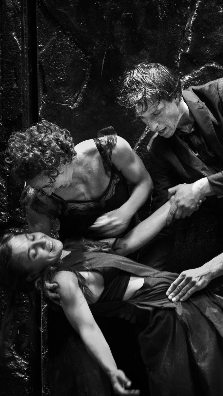 Five performers in dark clothing enact a dramatic scene on stage. The photo is in black and white.