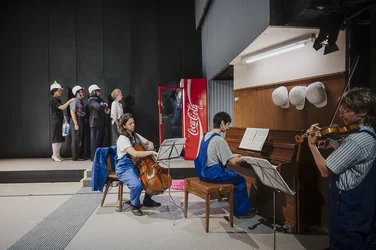 Three musicians play cello, piano and violin on stage. In the background, four performers wearing hard hats stand beside a red Coca-Cola vending machine.