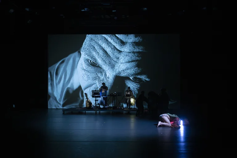 A stage performance with a large projected face in the background, while a person crouches on the floor and musicians are visible in the back.