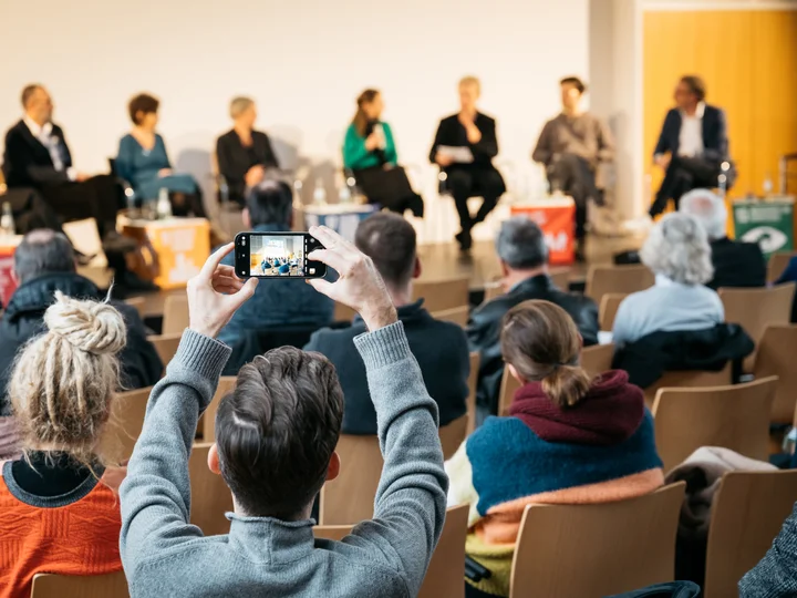 Gesprächsrunde auf einer Bühne, die von einer Person aus dem Publikum mit einem Handy fotografiert wird.