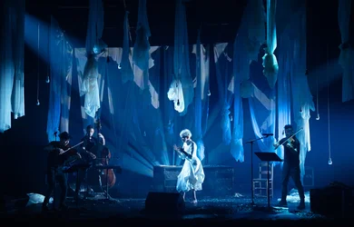 Auf einer Bühne stehen mehrere Musiker:innen mit Streichinstrumenten und Kontrabass. In der Mitte Joyce DiDonato in hellem Gewand. Von der Decke hängen zahlreiche halbtransparente Stoffbahnen, der Raum ist in blaues Licht getaucht.