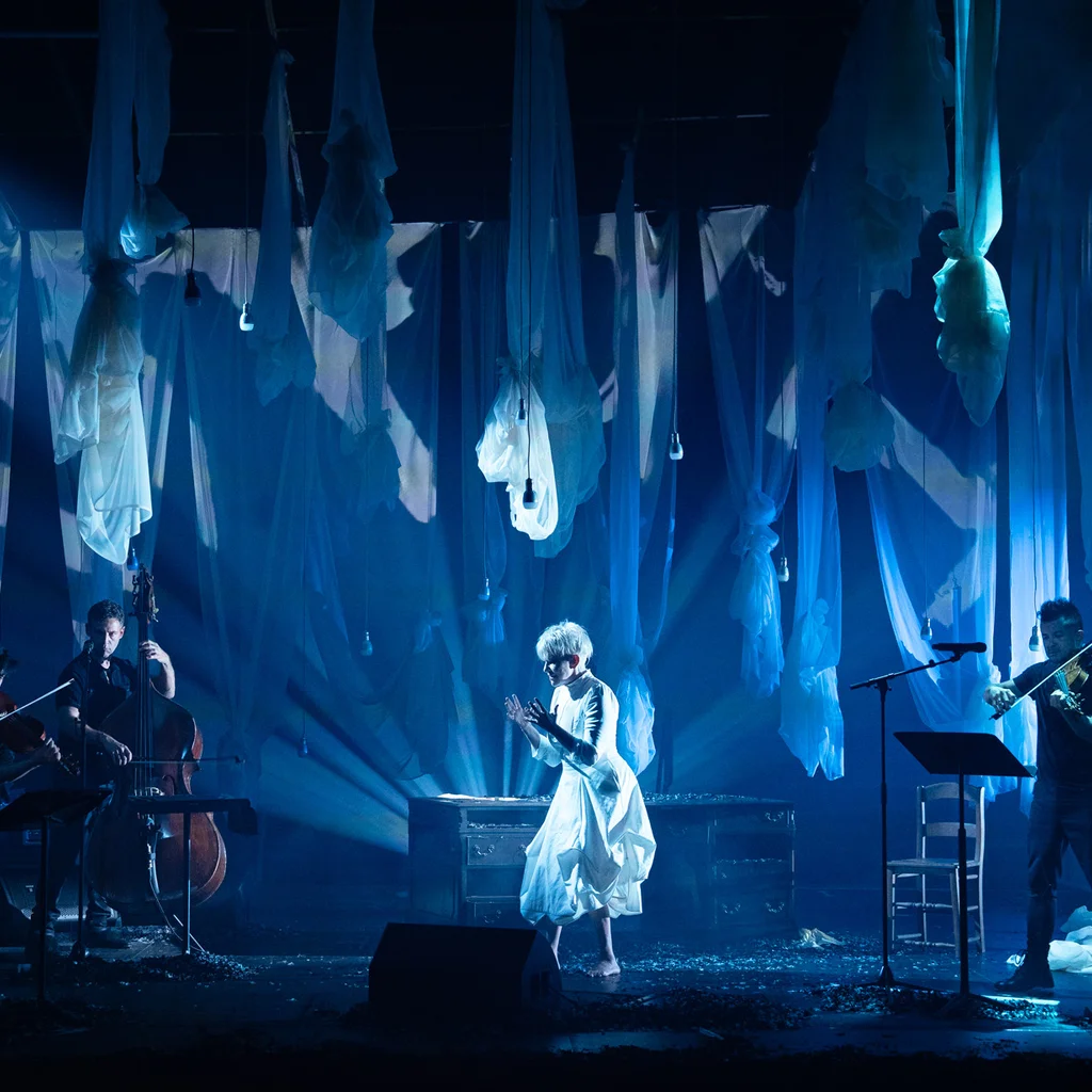 On a stage, several musicians with string instruments and a double bass are positioned. In the center, Joyce DiDonato in light-colored clothing stands in front of a table. Numerous semi-transparent fabric panels hang from the ceiling, and the space is lit in blue.