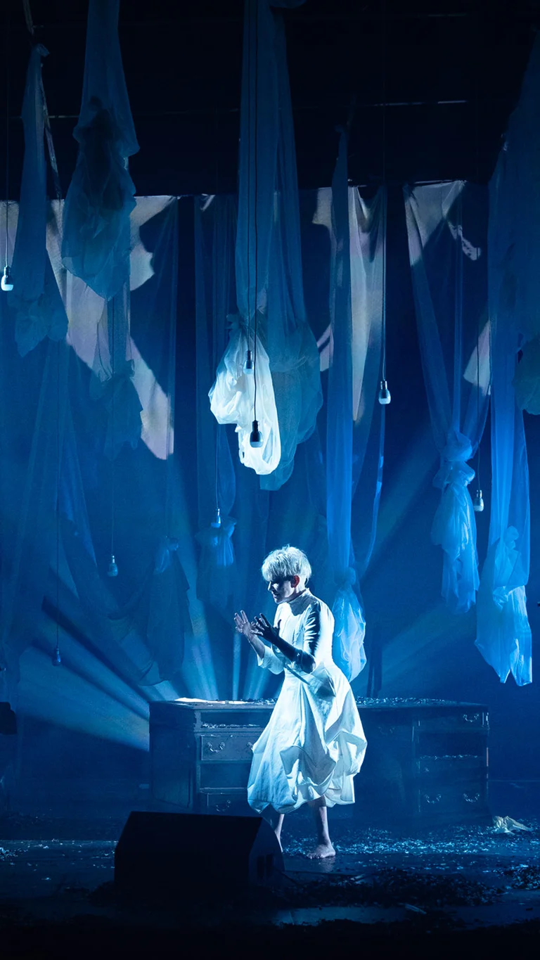 Auf einer Bühne stehen mehrere Musiker:innen mit Streichinstrumenten und Kontrabass. In der Mitte Joyce DiDonato in hellem Gewand. Von der Decke hängen zahlreiche halbtransparente Stoffbahnen, der Raum ist in blaues Licht getaucht.