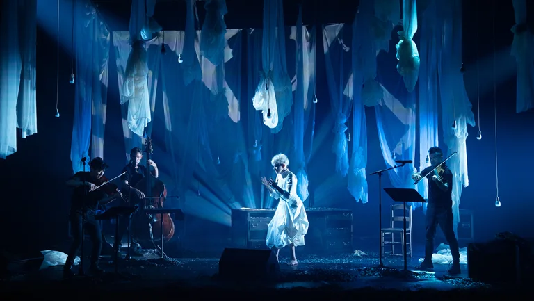 On a stage, several musicians with string instruments and a double bass are positioned. In the center, Joyce DiDonato in light-colored clothing stands in front of a table. Numerous semi-transparent fabric panels hang from the ceiling, and the space is lit in blue.