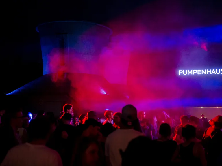 A group of people is celebrating at night in front of the "Pumpenhaus" during a Ruhrtriennale event. Red and blue fog fills the scene, while silhouettes of dancing attendees are visible in the foreground. The illuminated facade of the building contrasts sharply against the dark night sky.