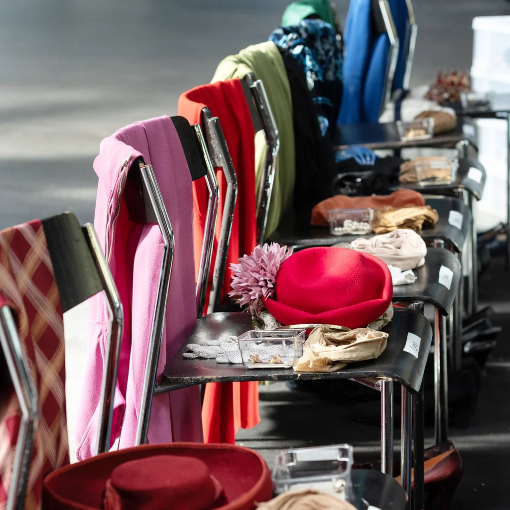Mehrere Stühle stehen in einer Reihe. Auf ihnen liegen farbige Kostümteile, Hüte, Schuhe und kleine beschriftete Behälter mit Accessoires für eine Produktion.