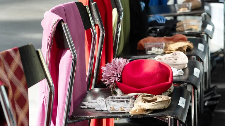 Mehrere Stühle stehen in einer Reihe. Auf ihnen liegen farbige Kostümteile, Hüte, Schuhe und kleine beschriftete Behälter mit Accessoires für eine Produktion.