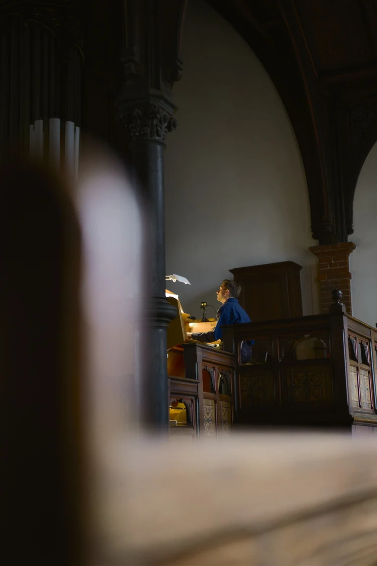 Organist James McVinnie at the organ in the Evangelical Church at the Market in Katernberg during the event 124 Years of Reverb. He is seated at the console on the gallery, illuminated by warm light, with the monumental organ pipes and historic wooden architecture in the background.
