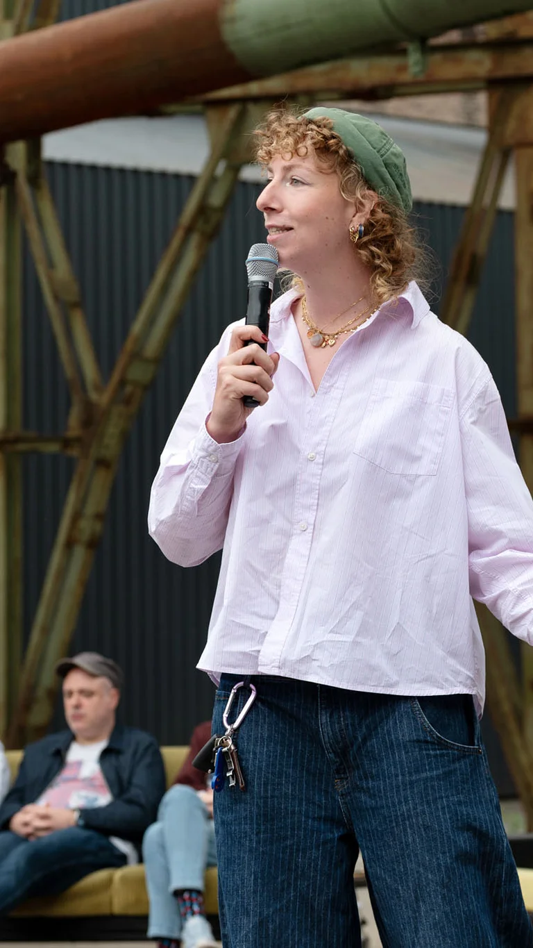 A person speaks with a microphone and book in hand in front of an audience; three people are seated behind on a sofa under industrial structures.