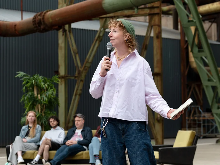 A person speaks with a microphone and book in hand in front of an audience; three people are seated behind on a sofa under industrial structures.