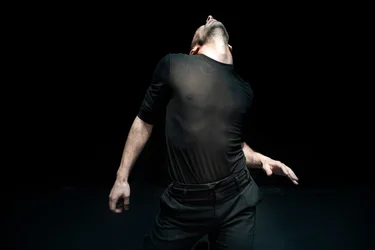 A dancer in black bends his upper body backward; his figure stands out in the light against the black stage background.