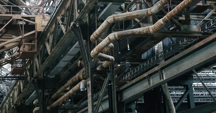 Nahaufnahme eines Netzwerks von verrosteten Rohren und Stahlträgern unter dem Dach der Jahrhunderthalle Bochum. Die komplexe Struktur zeigt die historische industrielle Architektur.