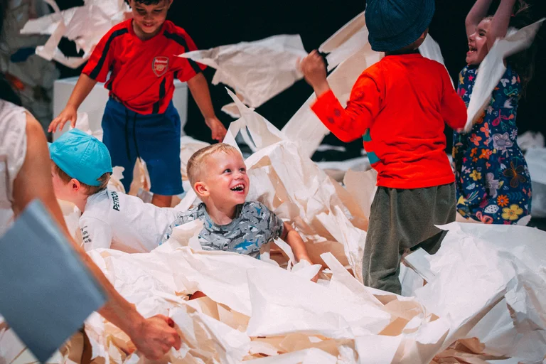 A group of children is playing excitedly with large sheets of paper. In the center, a blonde child wearing a gray T-shirt sits laughing in the paper, while other children around him toss the paper pieces into the air. A child in a red shirt and a blue hat raises their arms joyfully, while a girl in a colorful dress laughs beside them. In the background, a boy in a red jersey can be seen.