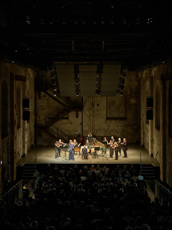 Blick aus dem Zuschauerraum auf Samuel Mariño mit Orchester auf der Bühne in industrieller Halle.