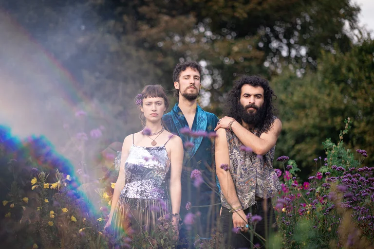 The band Karmakind stands in the middle of a blooming meadow with purple and yellow flowers. Three band members, one woman and two men, are visible. The scene is bathed in natural light, with a soft rainbow effect in the foreground.