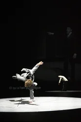 Solal Mariotte stützt sich mit einer Hand auf dem Boden ab und hebt die Beine seitlich an; im Hintergrund ist Anne Teresa De Keersmaeker auf der Bühne zu sehen.